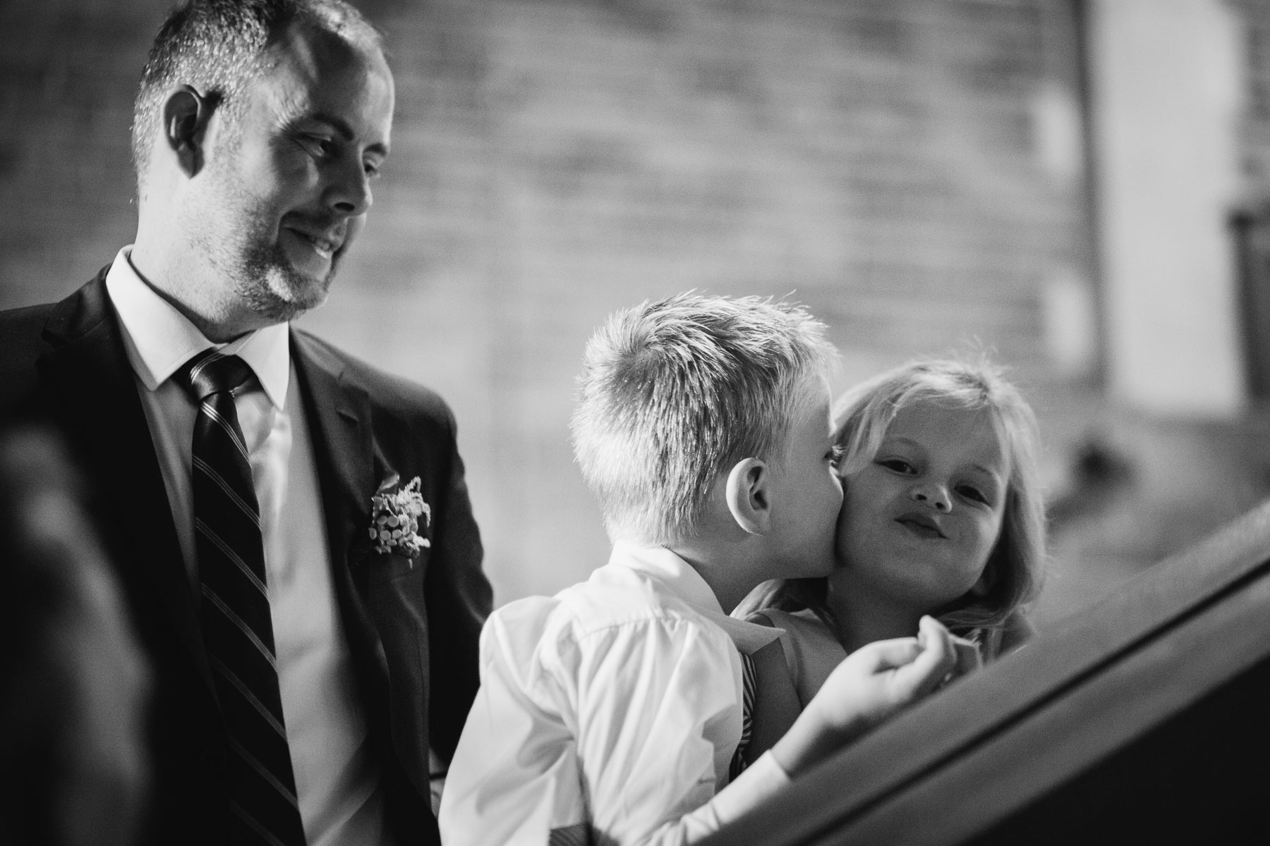 A boy kisses a girl on the cheek and she smiles towards the camera and the groom watches in this Hart House wedding photo.